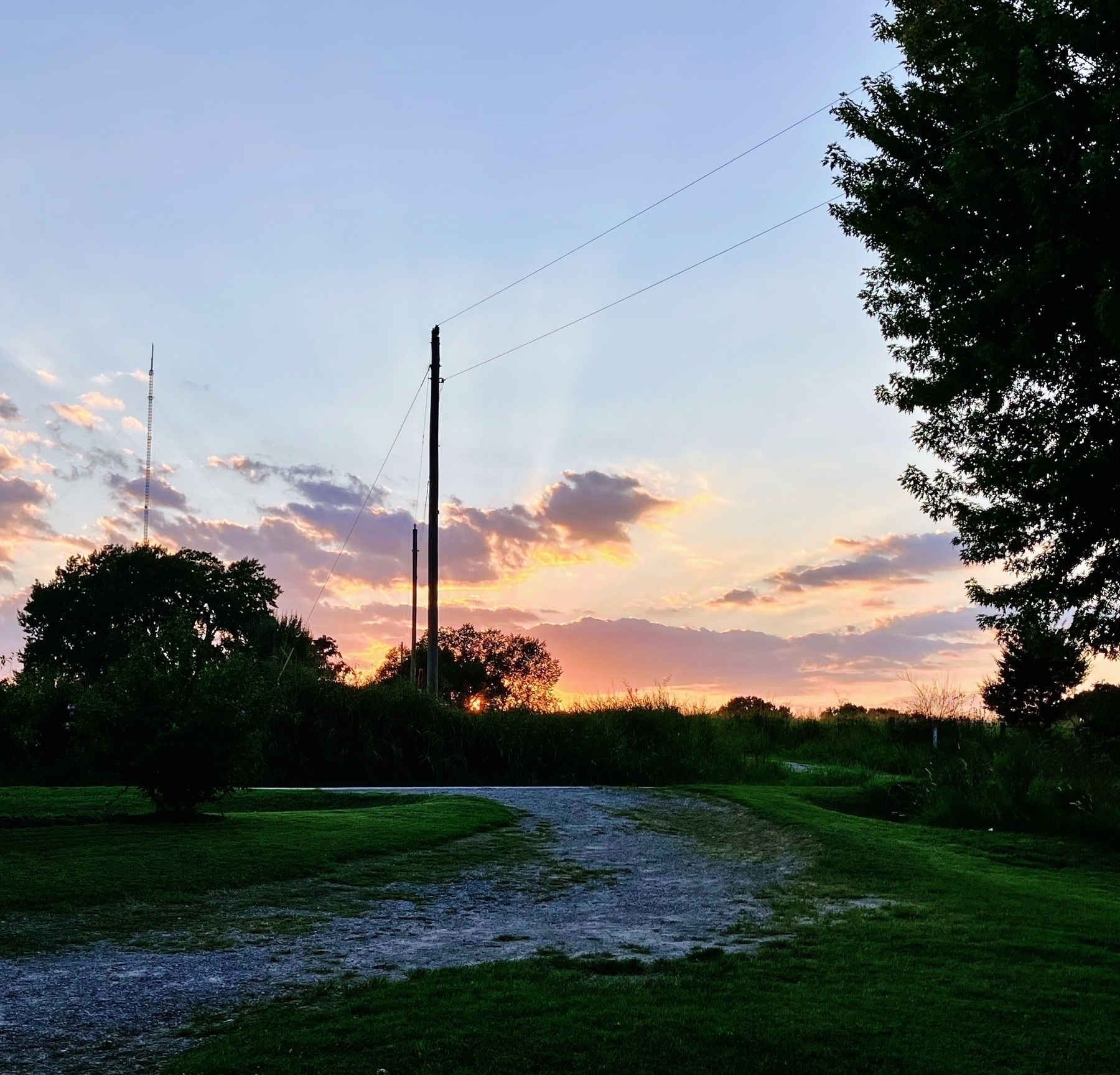 A gravel path curves through a grassy area at sunset. The sky is painted with shades of orange, pink, and purple, partially obscured by scattered clouds. Trees and bushes are silhouetted against the vibrant sky.