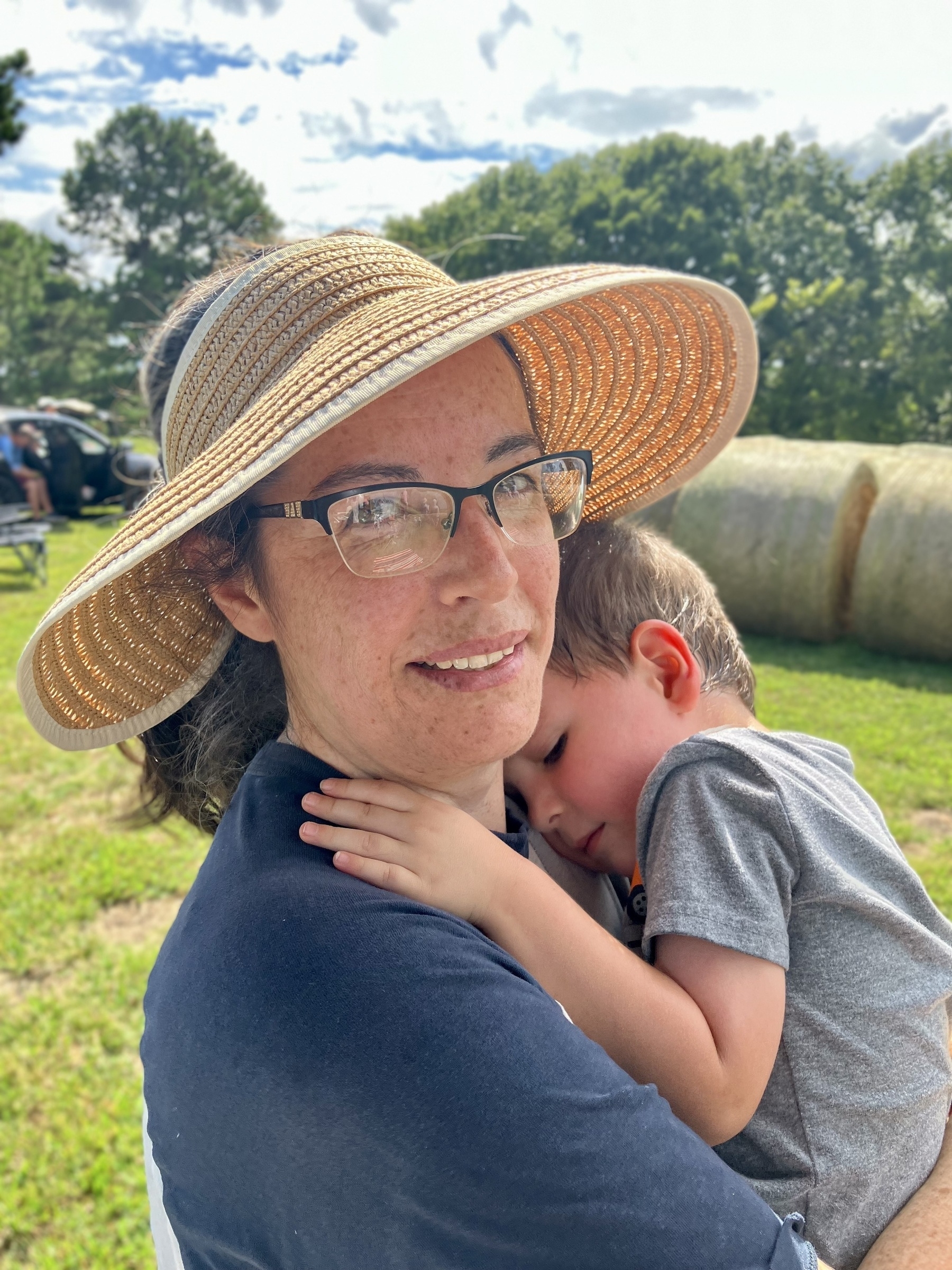A woman wearing a wide-brimmed straw hat and glasses is holding a young child who is resting against her shoulder. The background features a grassy area with hay bales and trees under a partly cloudy sky. The woman has a gentle expression as she looks at the camera.