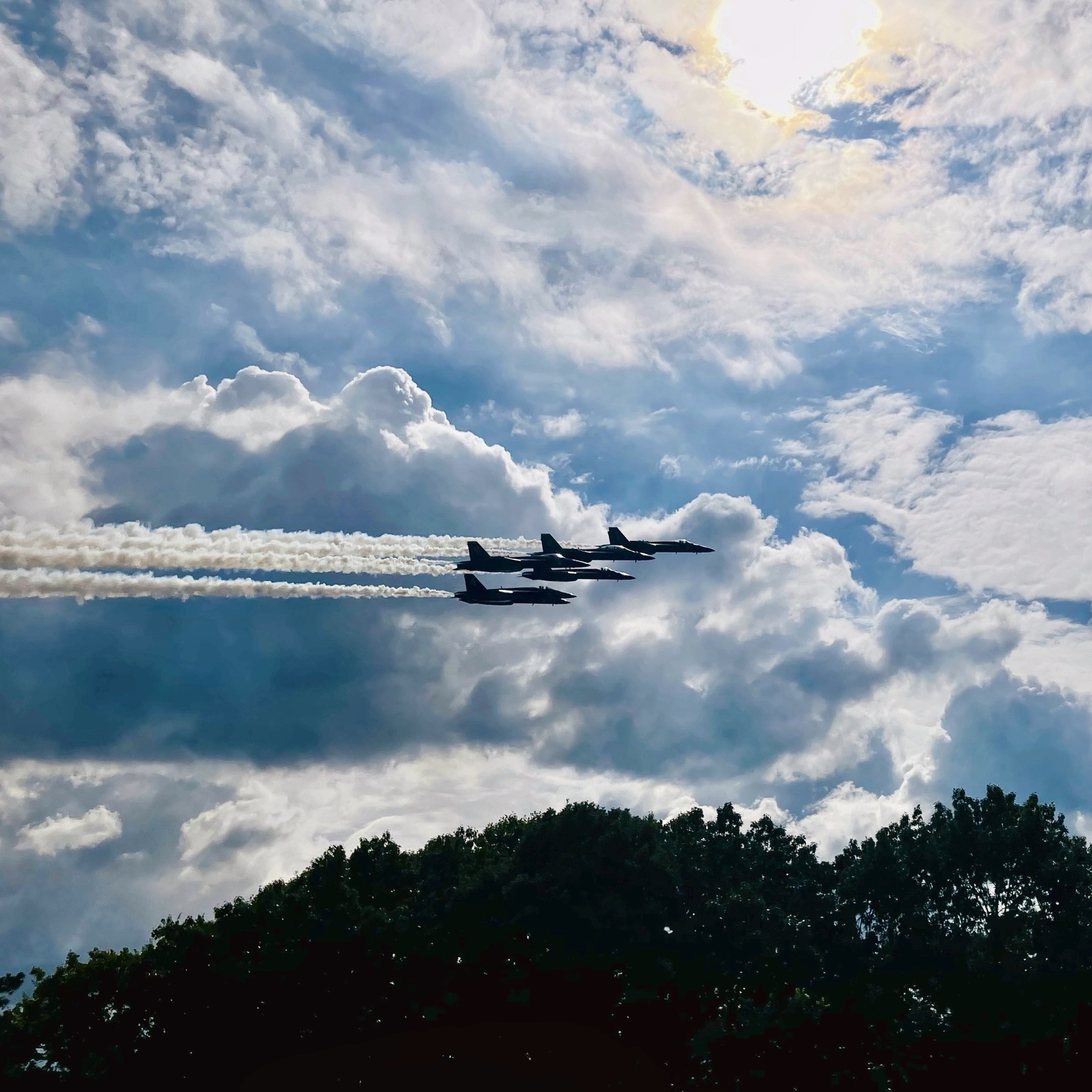 Auto-generated description: Four jets are flying in formation, leaving trails of smoke against a background of clouds and blue sky.