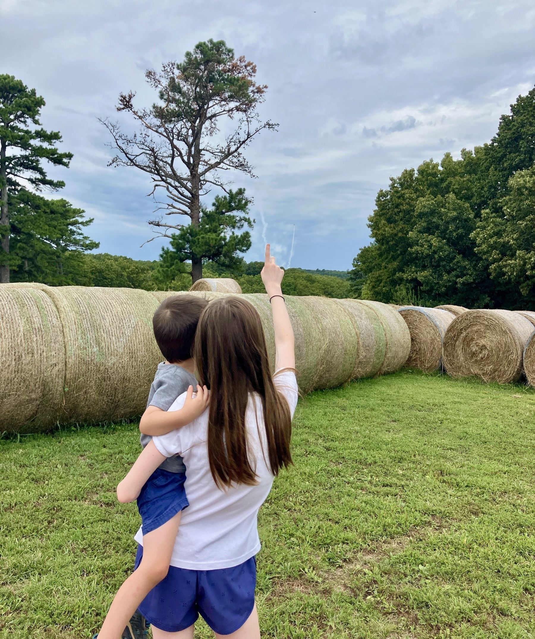 A girl stands on a grassy field, holding a small child on her hip. She is pointing towards the sky. Behind them are large, round hay bales and green trees under a cloudy sky.