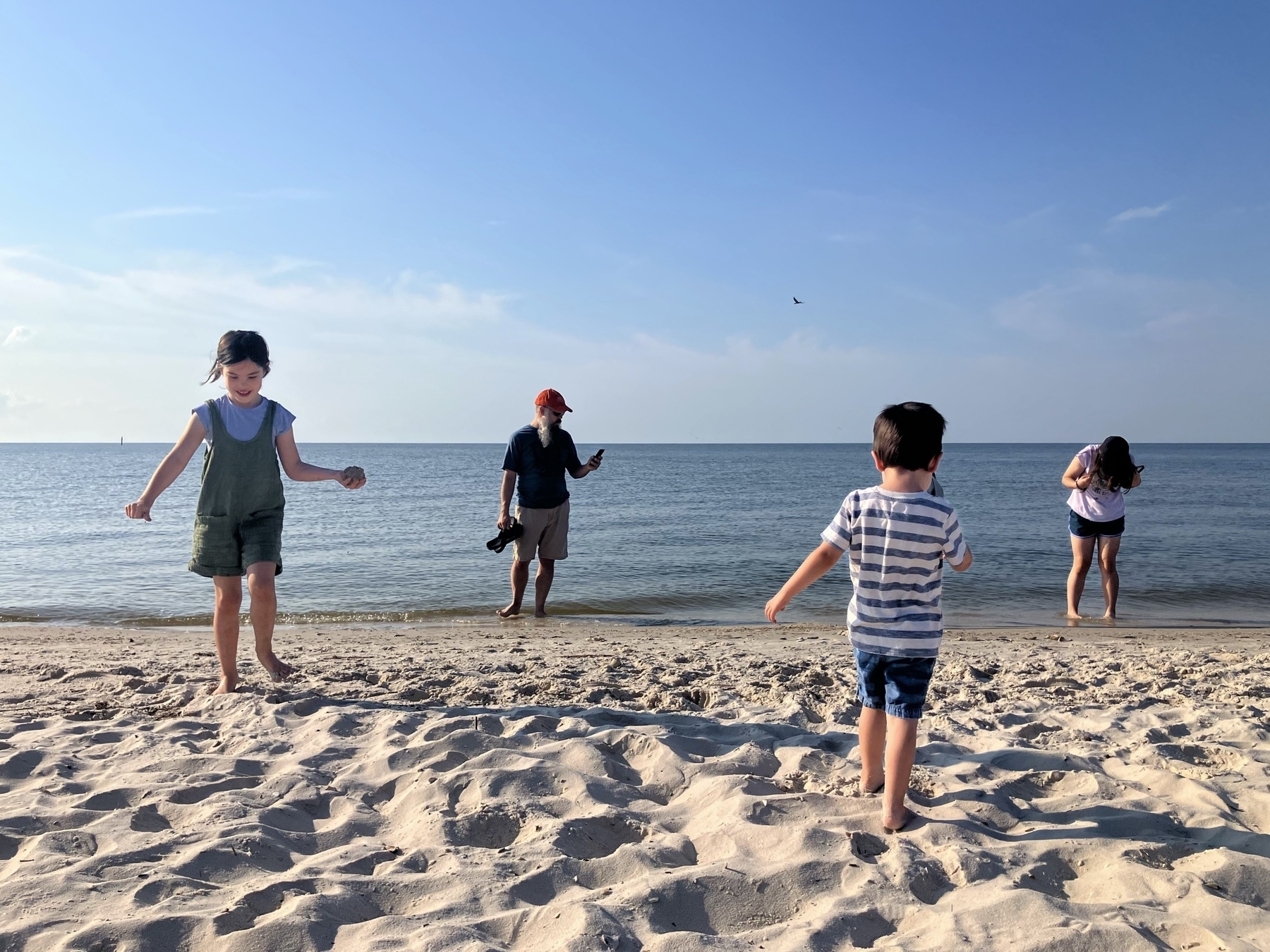 A group of people, including children, enjoy a sunny day at the beach, playing on the sand and near the water, with a clear blue sky and calm sea in the background.